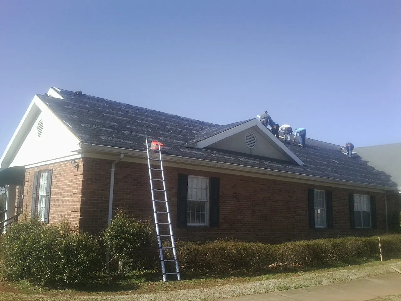 Crew tearing shingles off a brick ranch roof with a ladder set — Emergency Roof Repair work by our crew in Saint Stephen