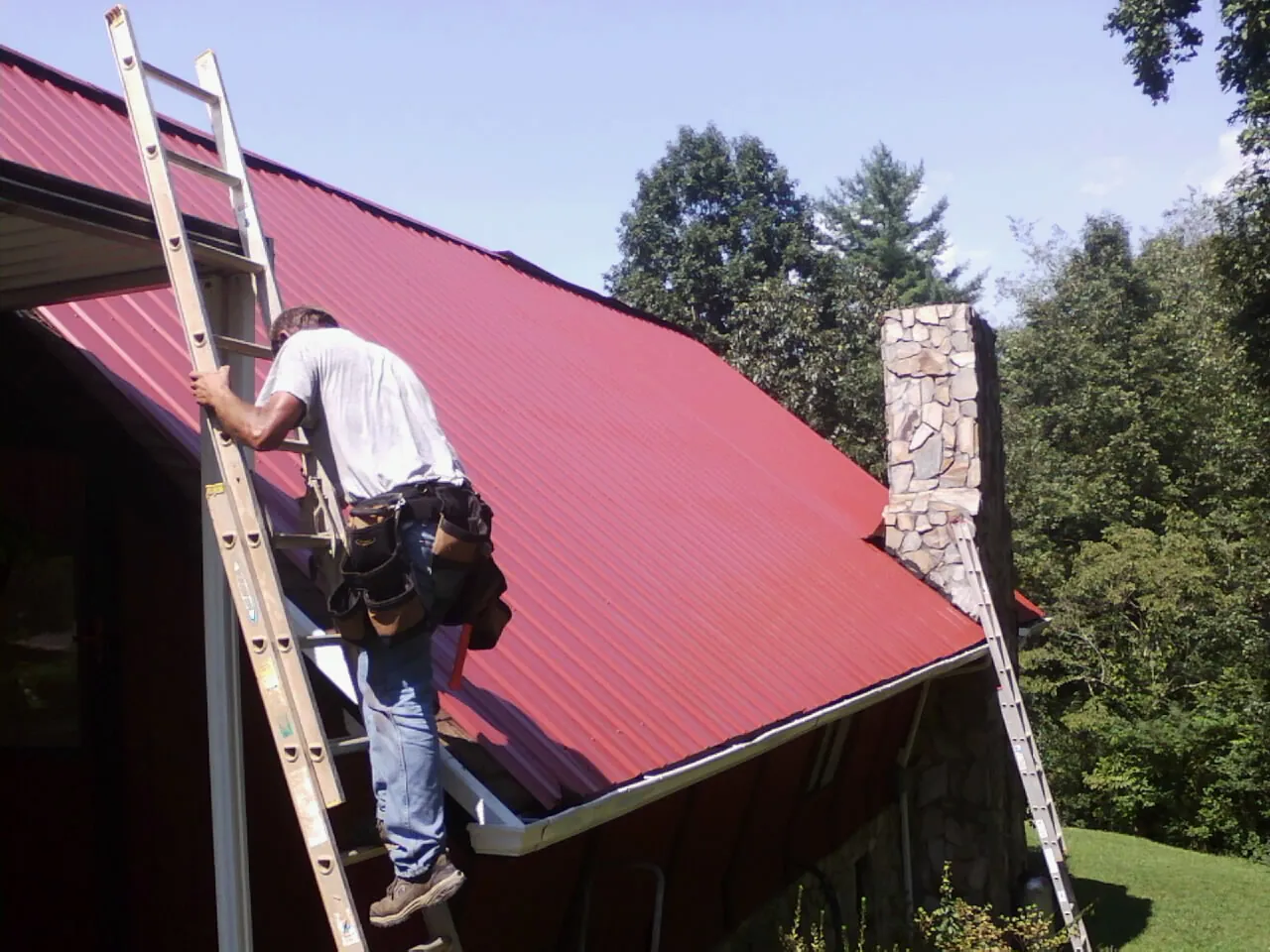 Roofer on a ladder beside a red metal roof and stone chimney — Soffit & Fascia Repair by Roofing Brothers in Saint Stephen